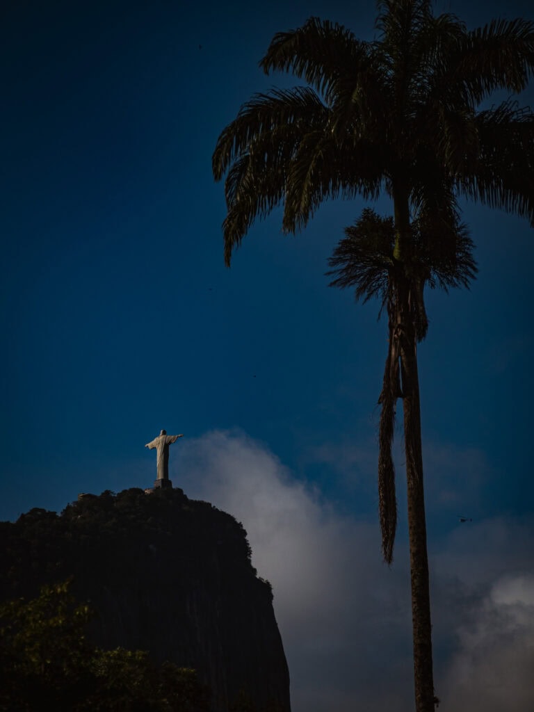 Christ the Redeemer statue in Rio de Janeiro at sunset with dramatic clouds and a tall palm tree in the foreground, Brazil travel photography
