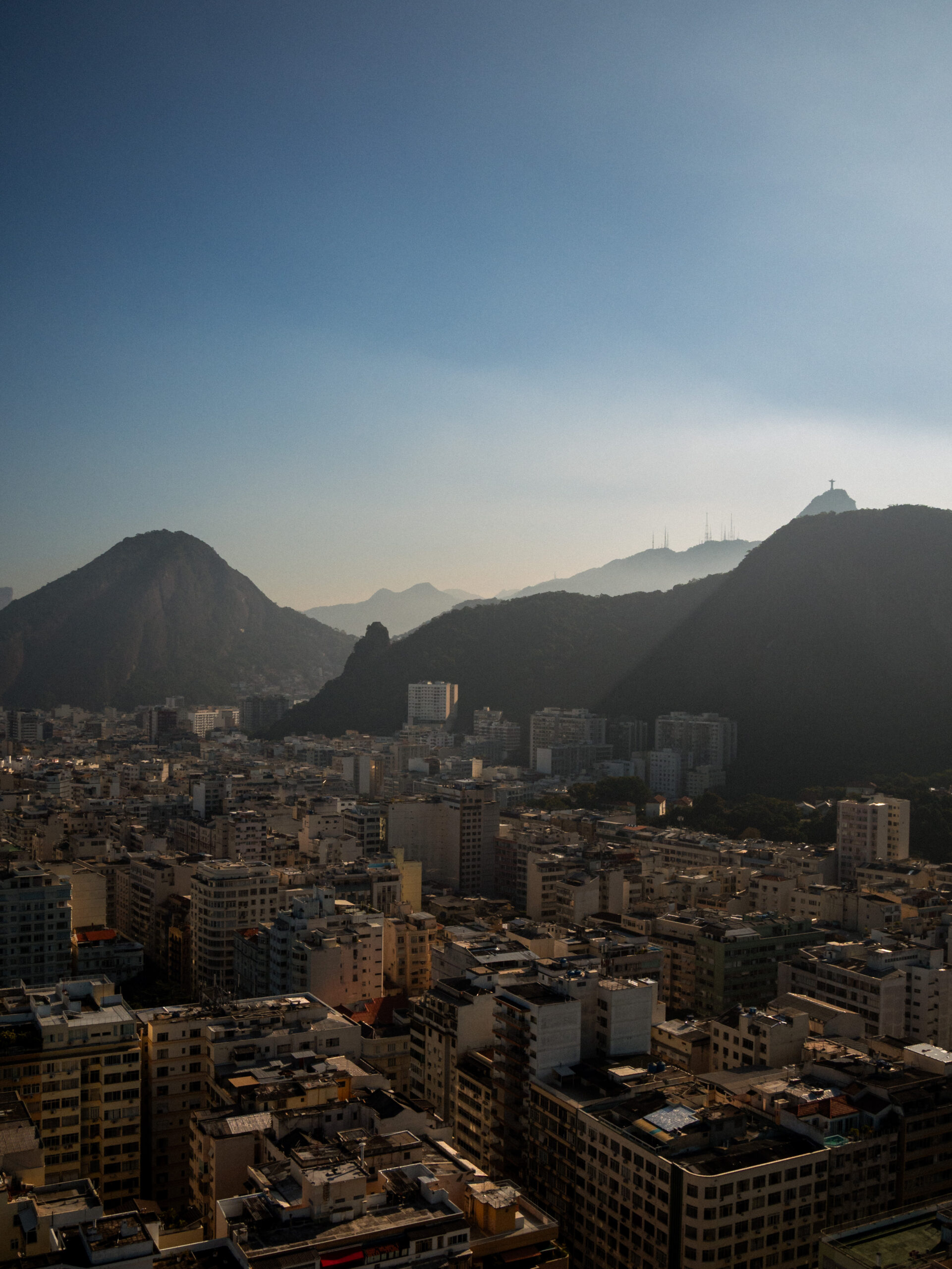 Aerial view of Rio de Janeiro city skyline with Christ the Redeemer visible in the distance, surrounded by layered mountain peaks at golden hour, captured with a cinematic tone from above