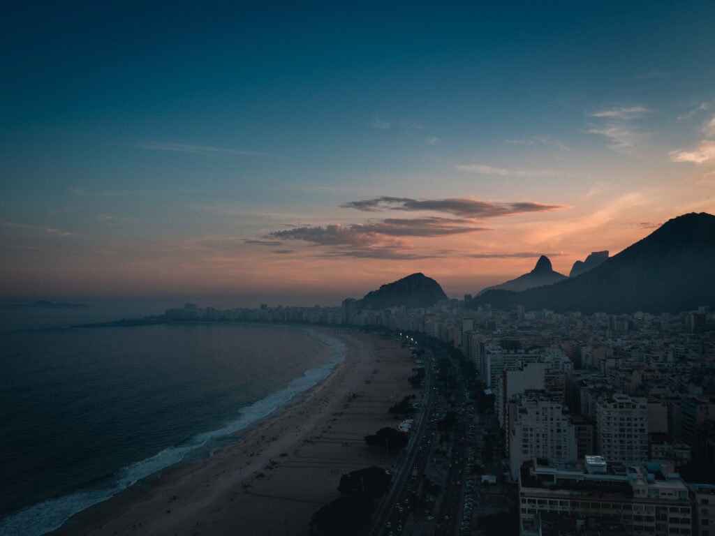 Aerial sunset view of Copacabana Beach in Rio de Janeiro with mountains in silhouette and city lights starting to glow along the coastline, captured during golden hour. A scenic highlight for Rio de Janeiro travel photography.