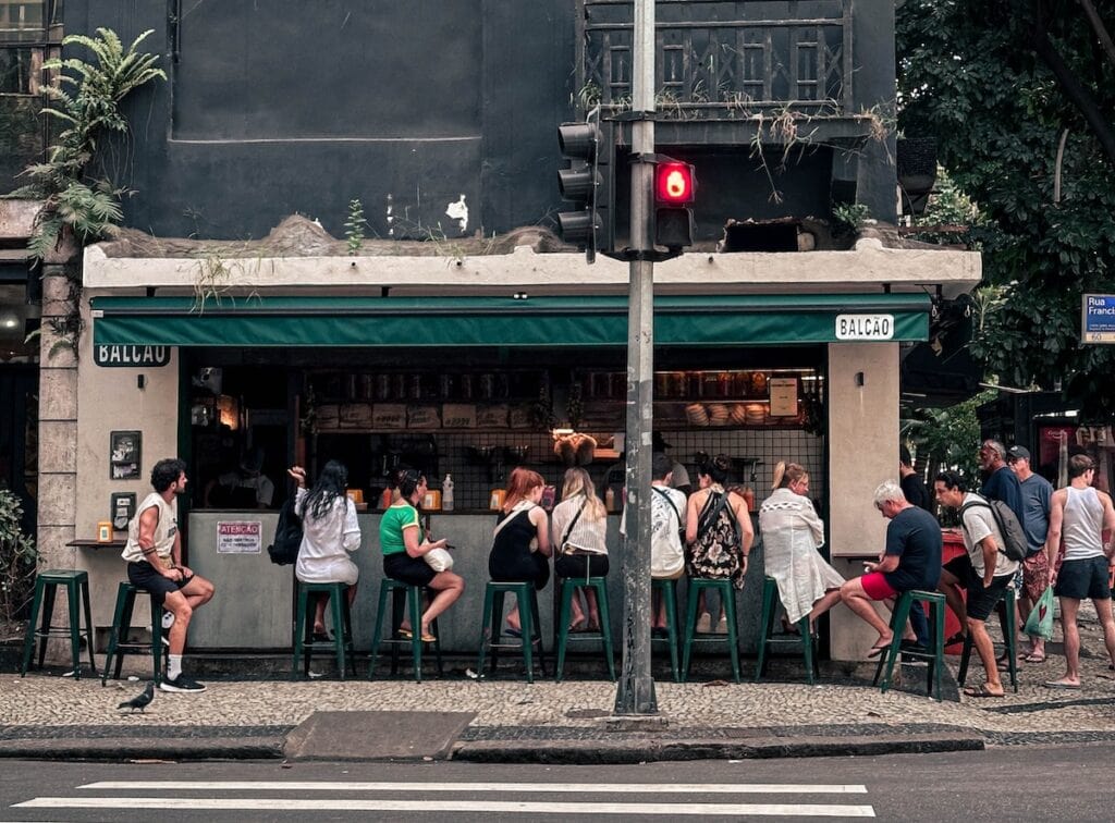 Locals and tourists sitting at an open-air Balcão restuarant in Ipanema, with a relaxed street scene capturing daily life in Rio
