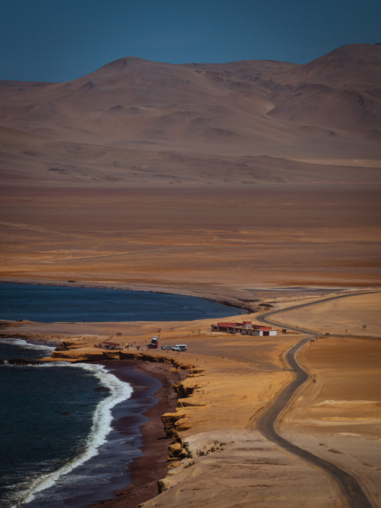 A high-angle photograph capturing the vast coastal desert landscape of the Paracas National Reserve in Peru. A winding dirt road leads down to a serene bay with deep blue water, framed by golden-brown cliffs and hills.