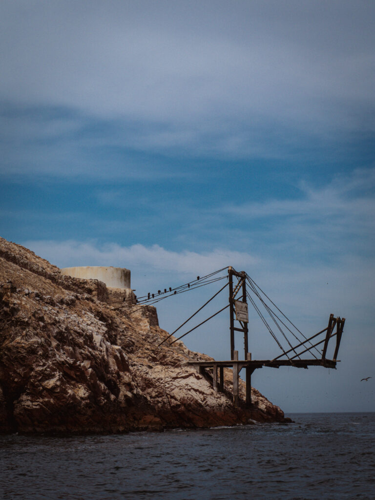 A photograph of an old, weathered guano loading crane and a small concrete structure on a rocky cliff face on the Islas Ballestas, Peru. A few birds are perched on the structure, which extends out over the choppy ocean.