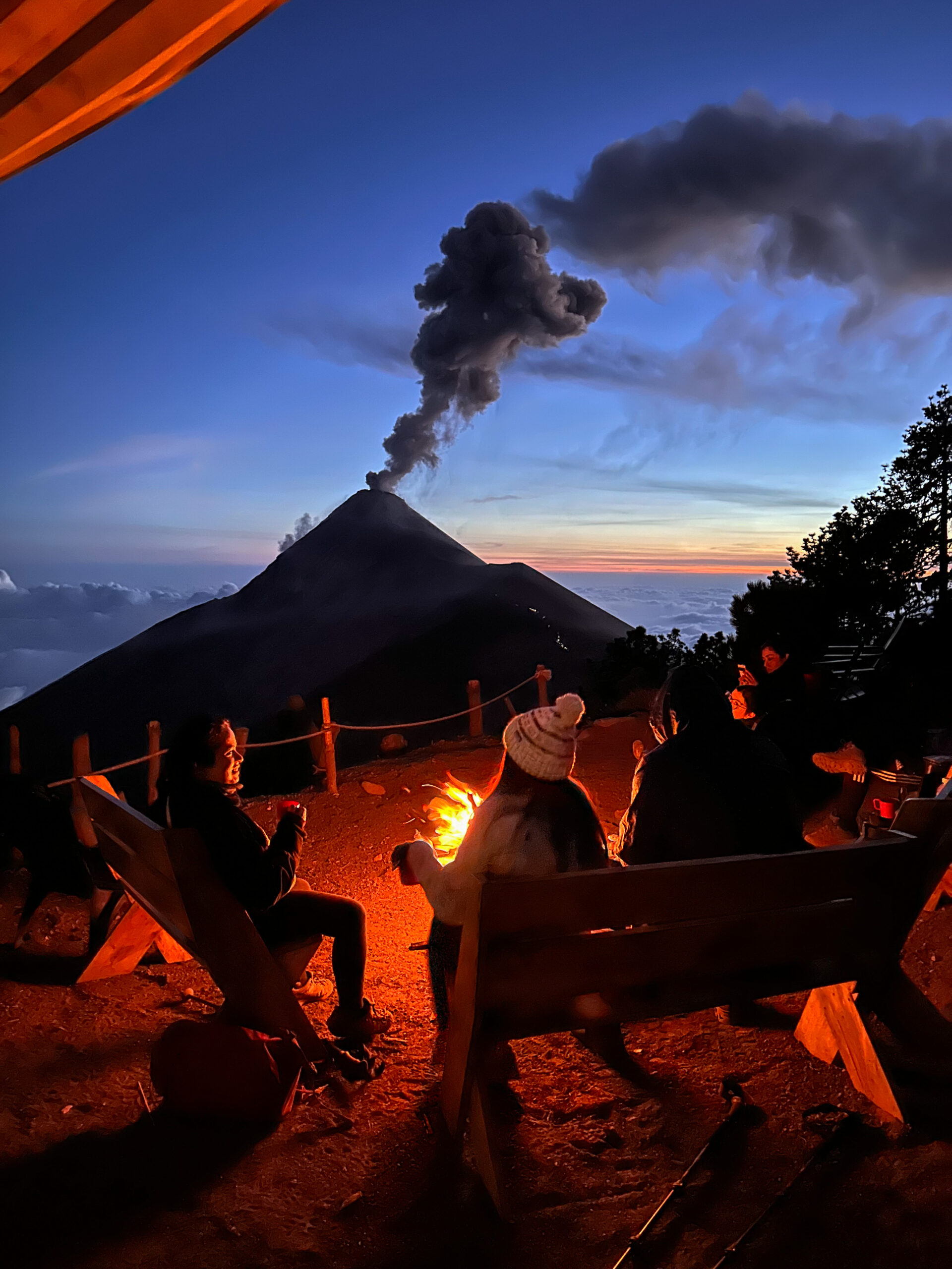 Campers sitting by a fire at Acatenango base camp with Volcán de Fuego erupting in the distance at dusk in Guatemala