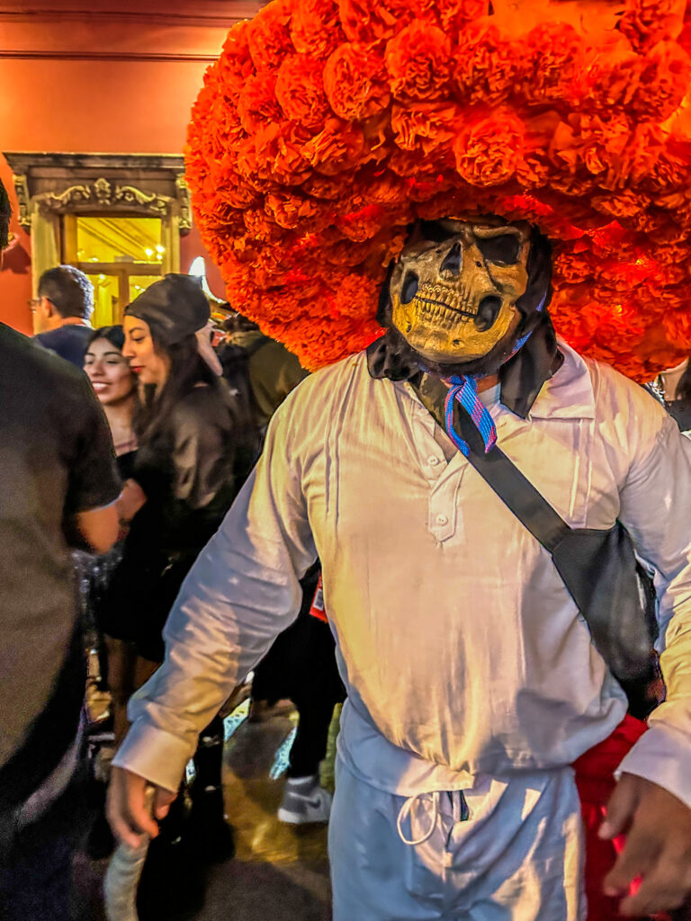 A man with a large orange marigold hat, skull mask and white clothes at Día de los Muertos in Oaxaca