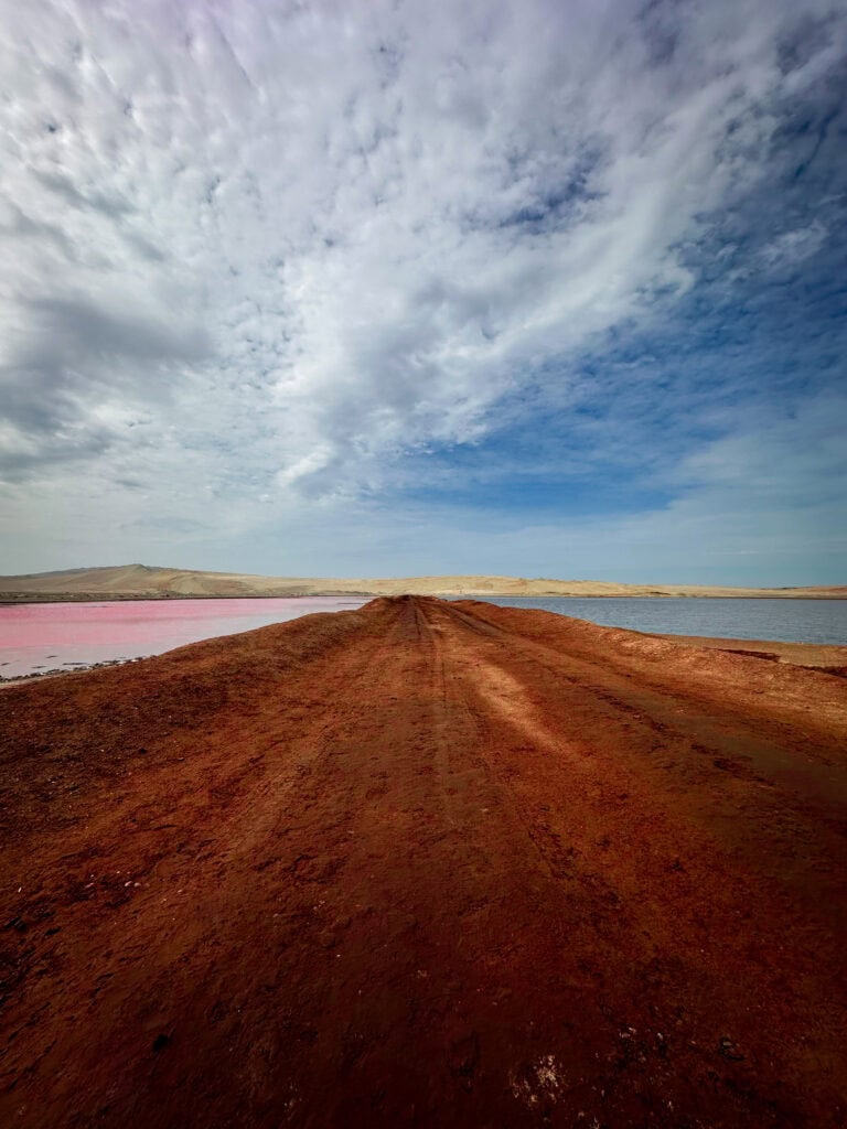 A ground-level view of a dirt track leading between two lakes in Paracas National Reserve, Peru. On the left, the water is a vibrant pink, while on the right, it is a deep blue, all set against a dramatic, cloudy sky.