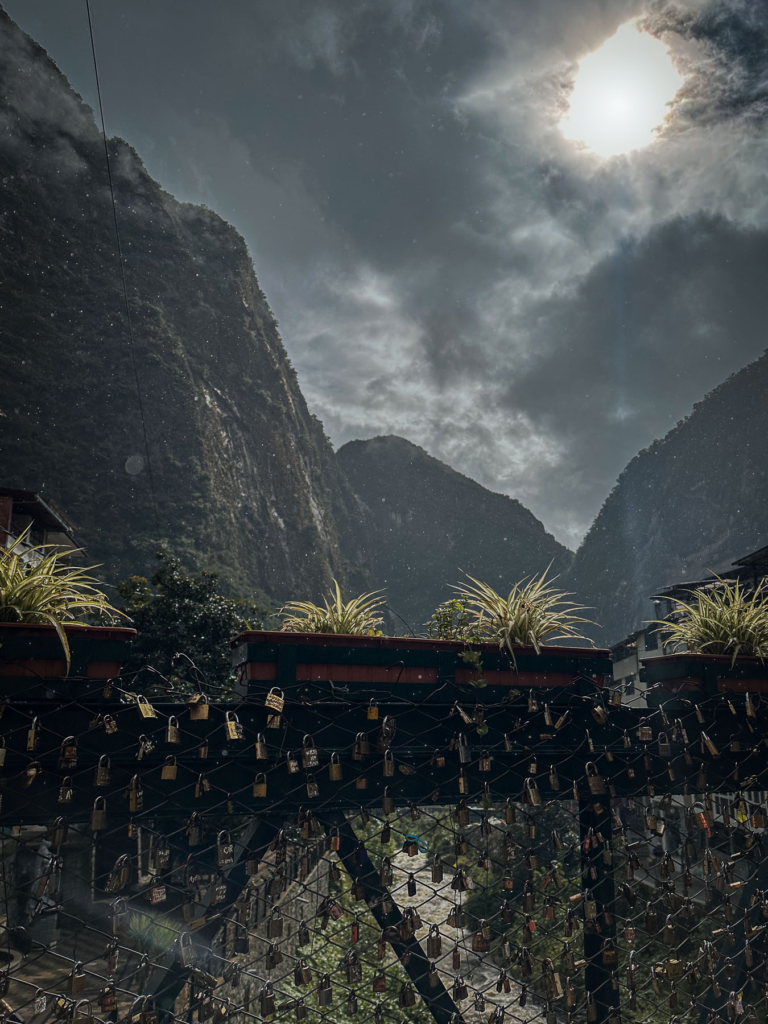 Love lock bridge in Aguas Calientes with dramatic mountain backdrop under cloudy sun